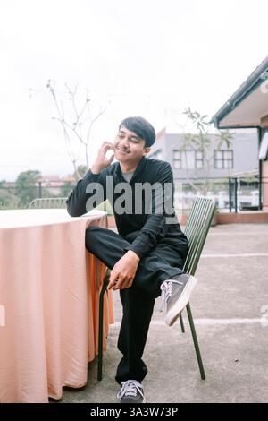 A young man sits at an outdoor table, lost in thought with a gentle smile. He appears relaxed and at peace, embodying a sense of quiet contentment and Stock Photo