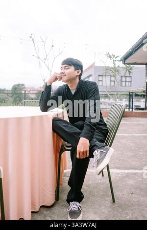 A thoughtful young man sitting relaxed outdoors on a chair next to a table. The Asian man is contemplating something with a pensive expression. The sc Stock Photo