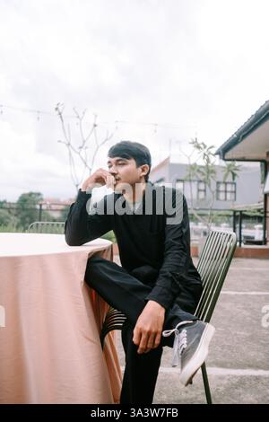 A thoughtful young man sits alone at a table on a rooftop patio, deeply contemplating. The serene outdoor setting provides a backdrop for his introspe Stock Photo