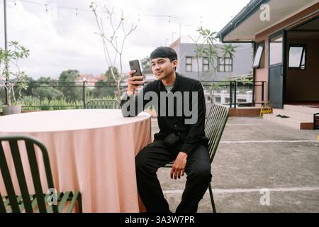 A young man is sitting at an outdoor café terrace enjoying his free time while using his smartphone. He might be chatting with friends, browsing socia Stock Photo