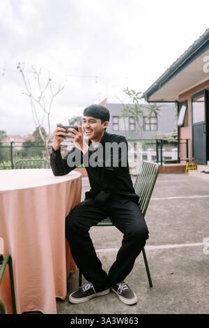 A young man sits on a patio, engrossed in playing a mobile game. With a happy smile, he holds his phone and looks captivated by the digital world in h Stock Photo