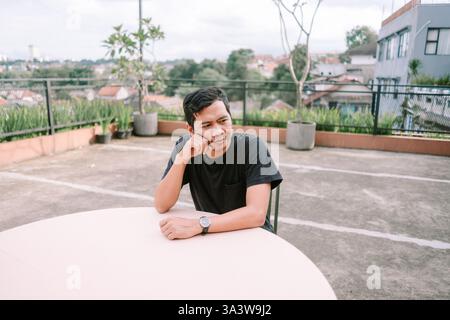 A young Asian man sits pensively at a table on a rooftop terrace, looking out at the cityscape. He appears thoughtful and relaxed, enjoying a moment o Stock Photo
