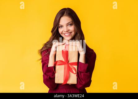 Young woman smiling while holding a wrapped gift in front of a yellow background Stock Photo