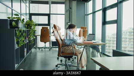 A modern professional woman working at a desk with a laptop in a stylish, bright office featuring large windows, plants, and contemporary furniture Stock Photo