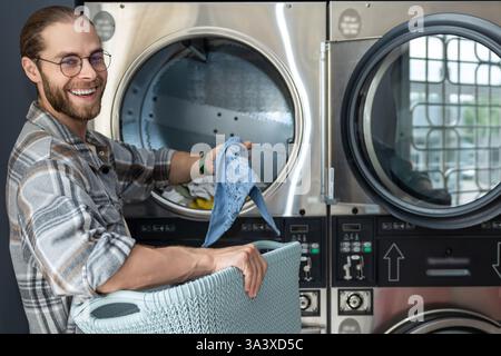 Man in public laundromat loading dirty clothes into washing machine Stock Photo