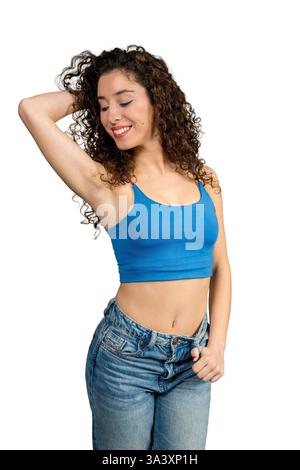 Young woman with curly hair, wearing jeans and blue top, is touching her hair and smiling Stock Photo