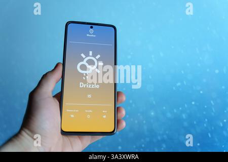 Hand holding a smartphone displaying a weather app with drizzle forecast. Raindrops falling in the blurred background. Weather technology and forecast Stock Photo