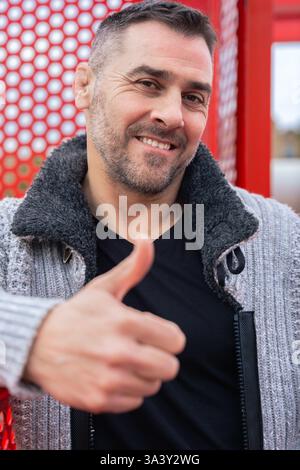 Confident Caucasian man with short dark hair giving a thumbs-up, smiling outdoors. Stock Photo