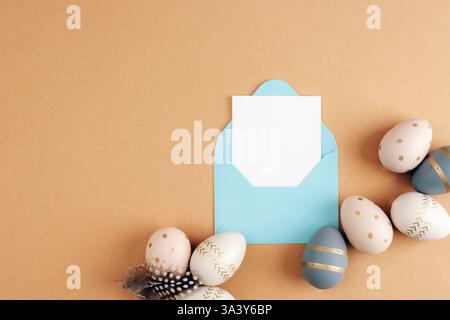 Minimalist Easter composition with a blue envelope, blank white card, and decorative eggs with golden patterns on a neutral brown background. Top view Stock Photo