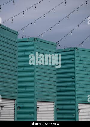 shipping containers painted turquoise standing on edge with white garage doors cut in the side light bulbs hanging from wires used for vendor booths Stock Photo