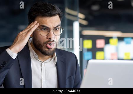 Upset suffering man at workplace inside office. Businessman unhappy and dissatisfied looking at laptop screen, reading online email with bad news. Stock Photo