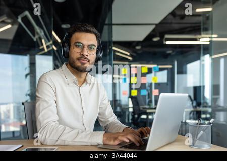 A focused young professional works on his laptop with headphones on in a modern office setting, displaying a thoughtful expression Stock Photo