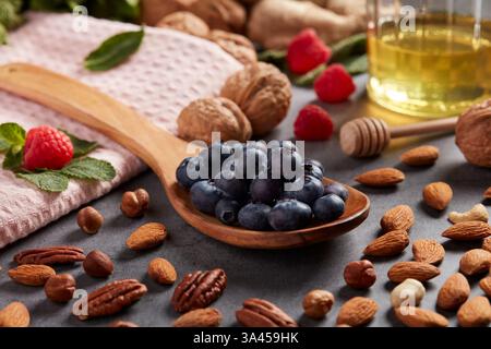 A wooden spoon full of blueberries sits on a gray surface scattered with walnuts, almonds, hazelnuts, raspberries, and a jar of honey. Stock Photo