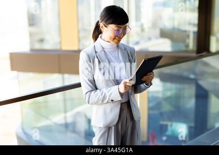 A focused Japanese business professional woman dressed in a stylish suit interacts with a digital tablet while standing near a large window. Sunlight Stock Photo