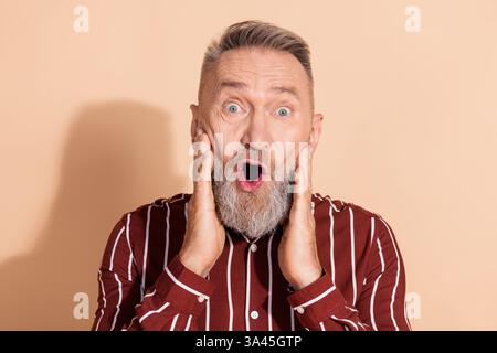 Elderly man in a striped shirt expressing astonishment with hands on face, against a beige background Stock Photo