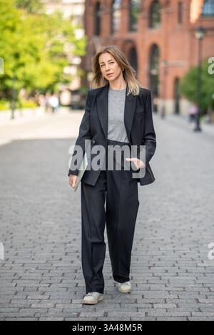 Stylish female entrepreneur with laptop stroll through urban streets, digital nomad and freelancer Stock Photo