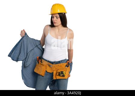 Young female construction worker wearing a yellow hard hat and tool belt, removing her denim shirt while looking down thoughtfully. She is dressed in Stock Photo