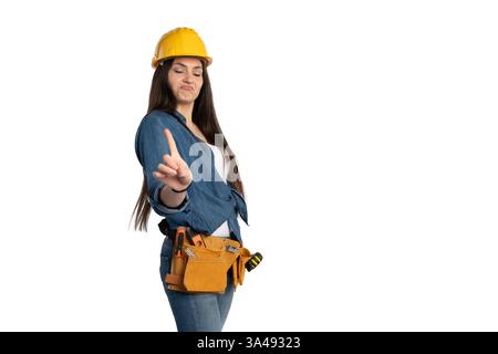 Young female construction worker wearing a yellow hard hat and tool belt, raising her finger in a disapproving gesture. She is dressed in a denim shir Stock Photo