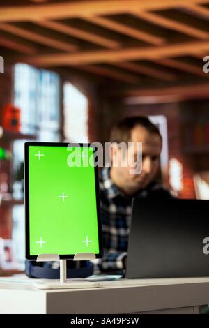 Focus on green screen tablet used by man in blurry background using mockup device, composing social media posts. Close up of chroma key tablet used by employee to monitor company social media Stock Photo
