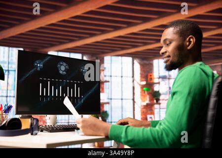 Smiling man in home office using financial application UI on PC screen showing company revenue and expenses. Accountant working from apartment doing annual profits review using computer display Stock Photo
