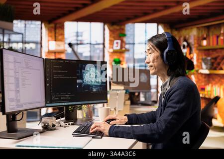 Senior programmer coding advanced AI algorithm in startup office, refining machine learning models to enhance automation, wearing headphones. Asian CTO manager listening music and coding Stock Photo
