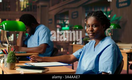 African american med school pupil studies pharmacology notes in a library, preparing for exams and advancing their healthcare career. Girl in scrubs works hard on medical school success. Camera B. Stock Photo