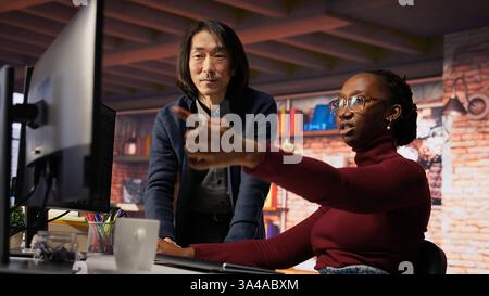 Man in AI startup office CTO position overseeing software engineer implementing machine learning. Artificial intelligence tech venture supervisor helps employee fix code bugs, camera B Stock Photo