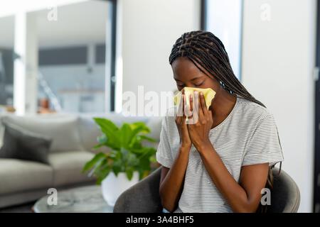 Woman sneezing into tissue at home, sitting on chair, feeling unwell, copy space Stock Photo