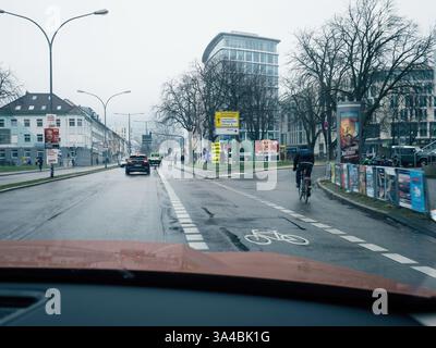 Freiburg, Germany - Feb 10, 2025: View from a car's dashboard as it travels a damp road in Freiburg, capturing a cyclist and urban scenery on a gloomy Stock Photo
