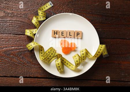 Bulimia. Squares with word Help, plate, cut carrot and measuring tape on wooden table, top view Stock Photo