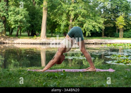 Young woman practicing downward-facing dog yoga pose on mat near tranquil lake surrounded by lush greenery. Concept of outdoor mindfulness Stock Photo
