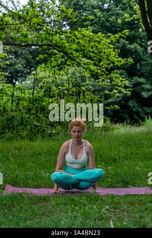 Young woman practicing seated yoga pose with crossed legs and hands on the ground in peaceful park. Concept of mindfulness, relaxation, and outdoor Stock Photo