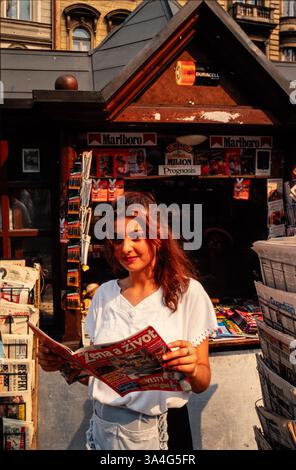 A young woman working at a street kiosk in Prague, Czechoslovakia, holding a magazine. The shop also sells cigarettes and batteries. Stock Photo