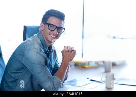 Smiling man in denim shirt sitting at desk in modern office environment, copy space Stock Photo
