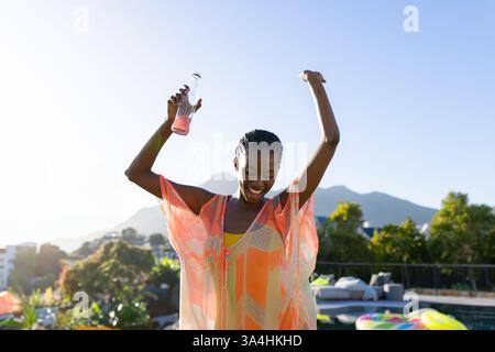 Joyful woman dancing by pool, holding drink, enjoying sunny day outdoors, copy space Stock Photo