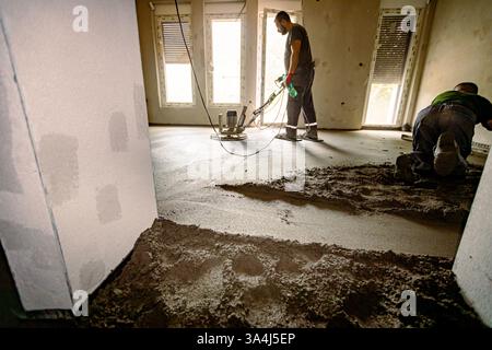 Construction workers are leveling semi-dry floor screed mixture over distribution heat system for floor heating, one manually and other one is using s Stock Photo