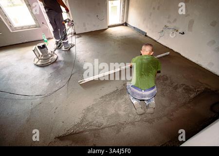Construction workers are leveling semi-dry floor screed mixture over distribution heat system for floor heating, one manually and other one is using s Stock Photo