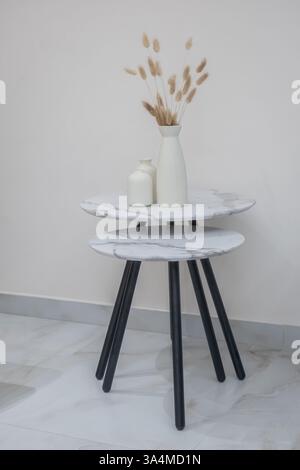 Two nesting tables with dry flowers in a vase in room. Set of 2 round modular coffee tables with black legs on light background. Nobody, selective foc Stock Photo