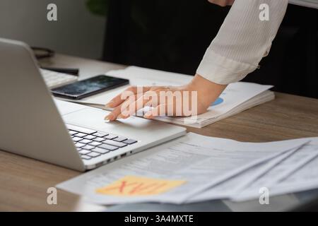 Document management for tax filing. Close-up of hands organizing financial paperwork with a laptop, depicting a busy workspace. Stock Photo