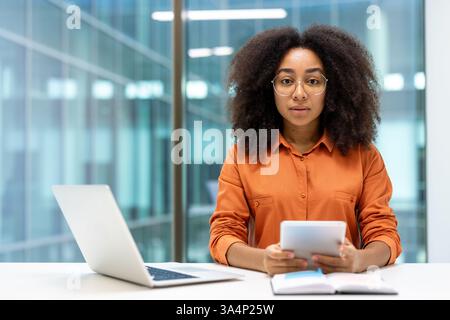 Portrait of a serious confident woman with a tablet computer in her hands. Businesswoman looks thoughtfully at the camera, works in the middle of the office, sits at a desk with a laptop. Stock Photo