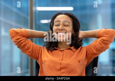 A young woman leans back in her office chair with hands behind her head, smiling and taking a moment to relax in a modern office space Stock Photo