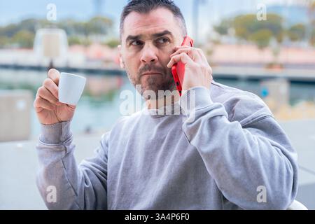 Caucasian man drinking coffee and talking on smartphone outdoors near the water. Stock Photo