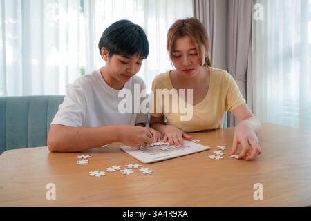 two asian friends working together on a puzzle at a wooden table, illustrating focus and teamwork. Stock Photo
