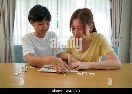 two asian friends working together on a puzzle at a wooden table, illustrating focus and teamwork. Stock Photo