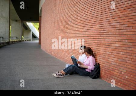 two asian college students seated on the ground, leaning against a brick wall. One holds an open book while the other uses a laptop, suggesting a coll Stock Photo