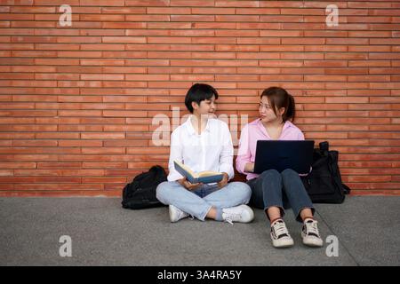 two asian college students seated on the ground, leaning against a brick wall. One holds an open book while the other uses a laptop, suggesting a coll Stock Photo