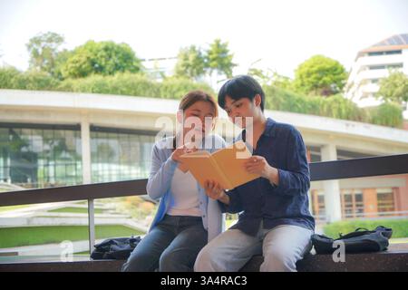 two asian college students seated on a bench in a modern university setting, engaging in a friendly discussion over a shared book Stock Photo