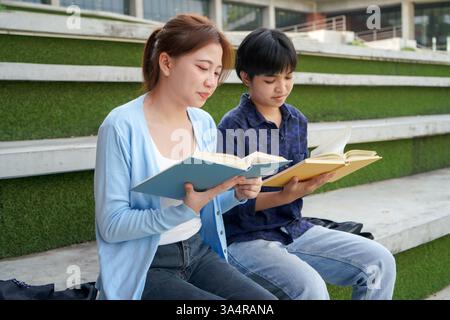 Relaxed scene of two asian college students seated on modern concrete steps with green turf, reading from colorful textbooks. Stock Photo