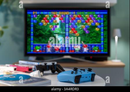 Milan, Lombardy, Italy - February 24, 2025: Light blue gamepad in focus in the foreground, retro Puzzle Bubble videogame displayed on a tv screen and Stock Photo