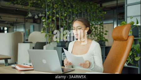A focused professional woman analyzing documents and working on a laptop in a modern, green office space with stylish furniture, productivity tools Stock Photo